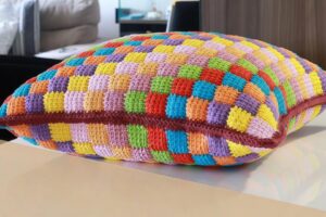 Detalhe de trabalho manual em crochê: Almofada quadrada em crochê com pontos baixos, fios de algodão coloridos em tons vibrantes, padrão xadrez e acabamento em ponto caranguejo bordô. — Produzido por Samuel Ramos - Blog do Crochê.