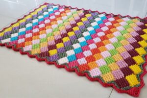 Detalhe de trabalho manual em crochê: Tapete de crochê com pontos baixos, fio de algodão colorido, padrão xadrez multicolorido, formato retangular com borda ondulada em vermelho. — Produzido por Samuel Ramos - Blog do Crochê.