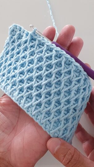 Amostra de crochê em tom azul claro utilizando o ponto trama dupla face, exibindo um padrão de relevo geométrico que lembra favos de mel. A peça é segurada por uma mão para mostrar o caimento e o detalhe do ponto.