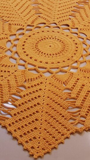 Foto em close de um tapete de crochê na cor mostarda com padrão que lembra folhas de coqueiro, destacando os detalhes dos pontos e a textura.